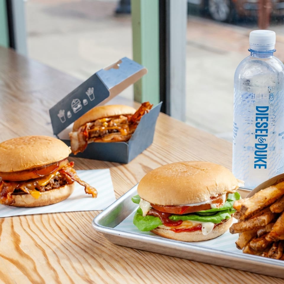 Three burgers with fries, a Coca-Cola bottle, and bottled water sit on a wooden restaurant table by a window.